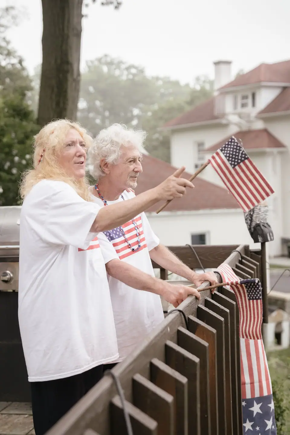 An older couple in white American flag print T-shirts waving small flags from a backyard deck