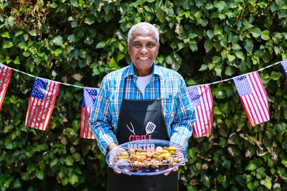 A smiling man in a plaid shirt and grill apron at a cookout under flag bunting